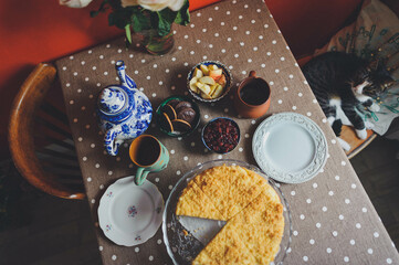 A table covered with a polka dot tablecloth with pastries, sweets, jam, tea and a cat lying on a chair next to it