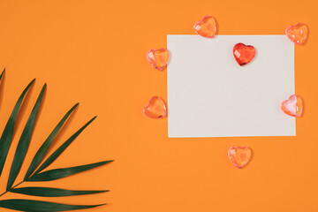 A white sheet of paper surrounded by hearts on an orange background with palm leaves. Minimal flat lay.