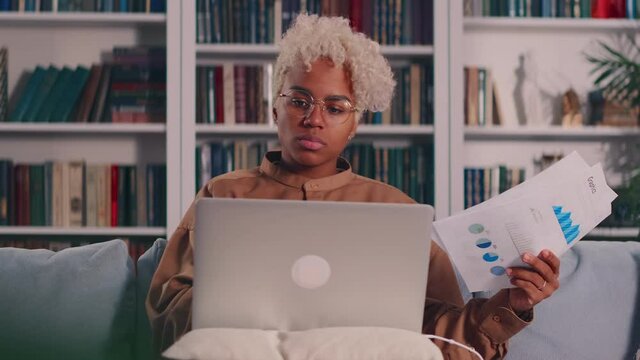 Serious young African American female sitting on sofa with laptop reading paper letters. Successful business woman working remotely at home, looking through documents, satisfied with financial reports