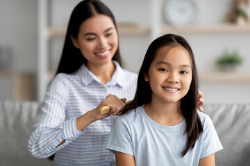 Caring asian mother brushing her cute daughter hair while sitting together on sofa at home in living room interior