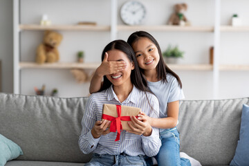 Pretty asian girl making birthday surprise for her mom and closing her mother eyes while sitting on couch