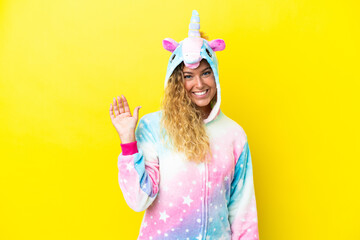 Girl with curly hair wearing a unicorn pajama isolated on yellow background saluting with hand with...