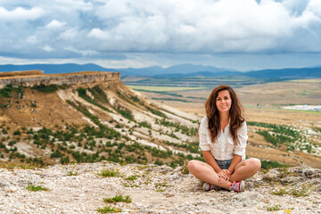 Fototapeta na wymiar A beautiful young woman in a white shirt is sitting on the edge of a cliff with an endless panorama, a White Rock in the Crimea. The concept of travel