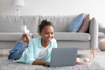 Happy young black girl student study at home with laptop, chatting with friends, watch video, lying on floor