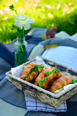 Basket of croissant sandwiches placed next to a vase with a white flower, set on a picnic blanket.