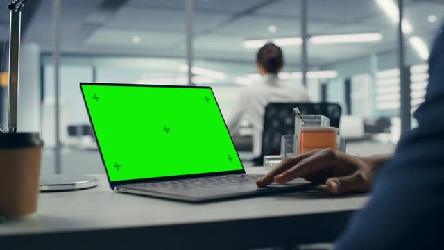 Successful Black Businessman Sitting at Desk Working on Green Screen Laptop Computer in Office. Anonymous Businessperson using Chroma Key Display. Stylish Bright Workplace. Over Shoulder Close-up