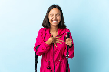 Young latin woman with backpack and trekking poles isolated on blue background smiling a lot