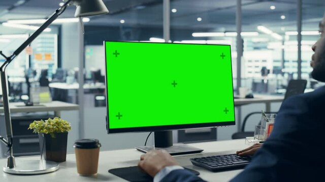 Successful Black Businessman Sitting at Desk Working on Green Screen Laptop Computer in Office. African American Businessperson using Chroma Key Display. Over Shoulder Arc Shot