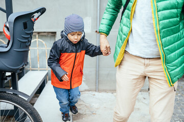 Unrecognizable european father in green jacket taking his son after school or kindergarten at home with bicycle. Happy family concept.