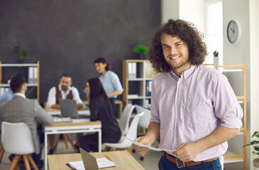 Happy handsome curly young man in casual shirt standing in modern office workplace, holding document, smiling and looking at camera. Portrait of successful confident business professional at work