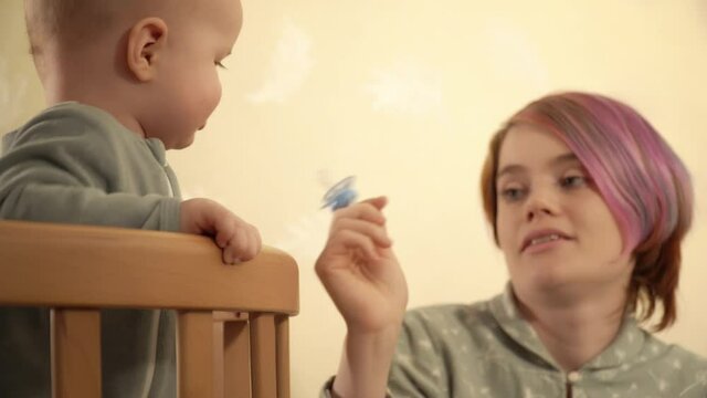 Mother is giving a pacifier to her son who is standing in the crib and ridding it. Young caucasian woman is trying to calm her baby by a soother. She is looking with love at her kid. Motherhood theme.