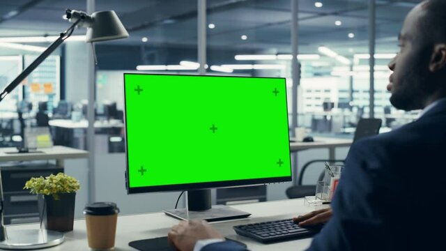 Successful Black Businessman Sitting at Desk Working on Green Screen Laptop Computer in Office. African American Businessperson using Chroma Key Display. Over Shoulder Zoom In