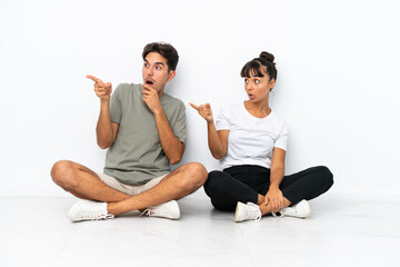 Fototapeta na wymiar Young mixed race couple sitting on the floor isolated on white background pointing finger to the side with a surprised face