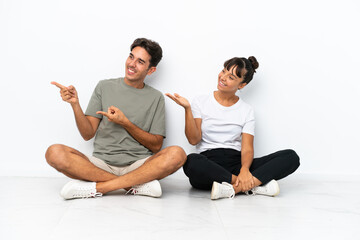 Young mixed race couple sitting on the floor isolated on white background pointing back and presenting a product