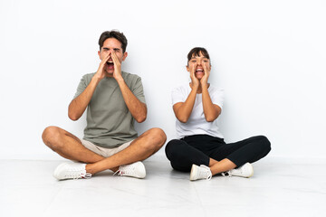 Obraz premium Young mixed race couple sitting on the floor isolated on white background shouting with mouth wide open to the lateral