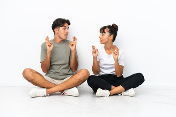 Fototapeta na wymiar Young mixed race couple sitting on the floor isolated on white background with fingers crossing and wishing the best