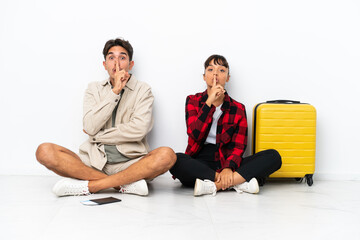 Young mixed race travelers couple sitting on the floor isolated on white background showing a sign of silence gesture putting finger in mouth