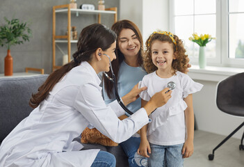 Home visit of a pediatrician. Friendly female doctor applies a stethoscope to listen to the heartbeat of a little cute girl. Concept of child health and pediatric medical examination.