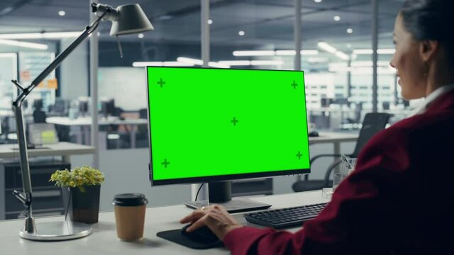 Successful Caucasian Businesswoman Sitting at Desk Working on Green Screen Laptop Computer in Office. Beautiful Businessperson using Chroma Key Display. Over Shoulder Static Shot