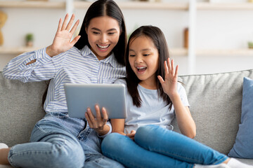 Happy asian mother and daughter having video call on digital tablet, sitting on sofa and waving at pad screen