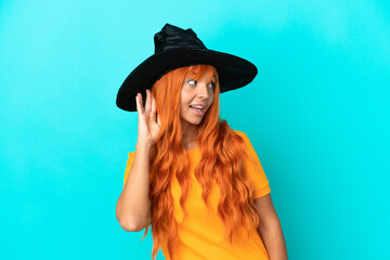 Young woman disguised as witch isolated on blue background listening to something by putting hand on the ear