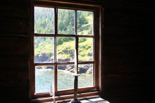 A candle in an antique candle holder and a quill pen in an ink well, on an old wooden desk in front of a window. View of coastline through window.
