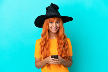 Young woman disguised as witch isolated on blue background sending a message with the mobile