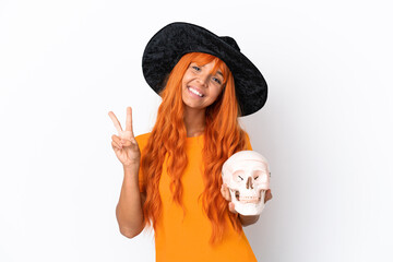 Young mixed race woman disguised as witch holding a skull isolated on white background smiling and showing victory sign