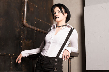 Stylish black haired girl with black makeup with suspenders and hat at the steel background and pipes