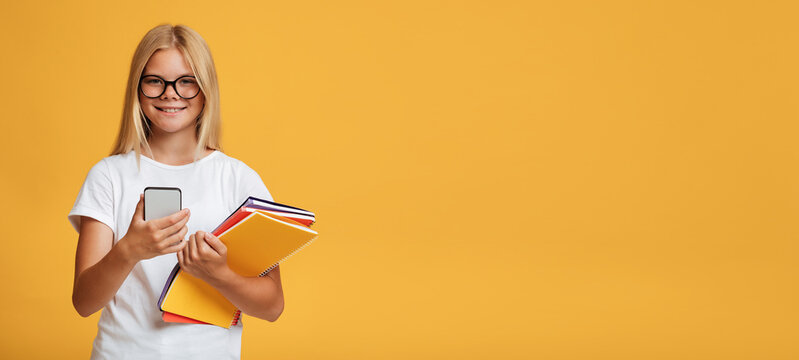 Cheerful cute teenage caucasian blonde girl in glasses with notebooks uses phone for study
