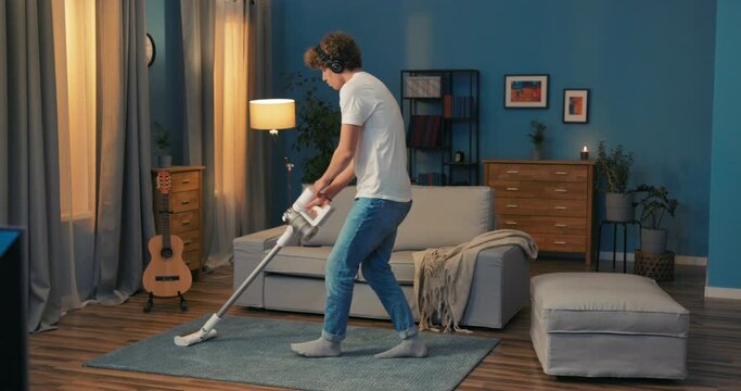 Handsome boy with curls on head does household chores vacuums the living room carpet with wireless headphones on ears listens to music dances with hand in pocket, passes the time
