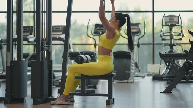 Hispanic woman sitting on a simulator in the gym pulls a metal rope with the weight pumps up the muscles of the back. brunette woman pulls on simulator. performing exercise for back muscles simulator