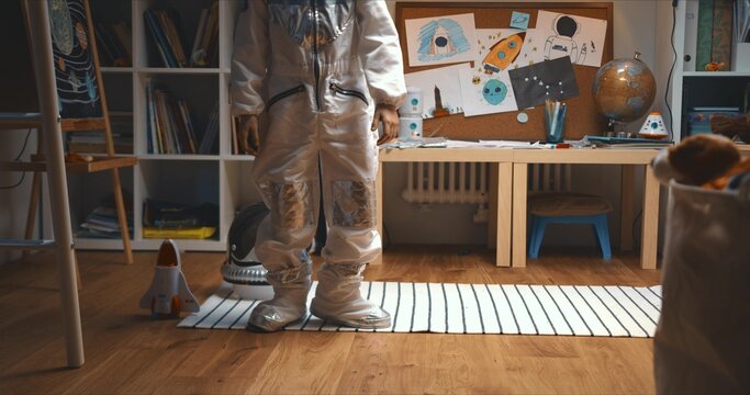 Childhood Dreams. Happy Cute Little Boy Wearing Astronaut Suit Sitting Down In Room To Play With Rocket Space Ship Toy.