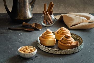 Sweet cinnabon buns lie on a ceramic plate, next to a saucepan with brown sugar, cinnamon sticks in a glass glass, a wooden scoop and spoon and a coffee pot. Homemade cakes in the shape of a snail.
