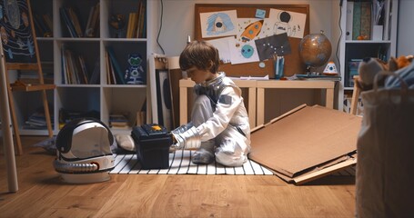 Childhood activities. Little boy in astronaut suit enters room with big cardboard piece, tool box to build space ship.