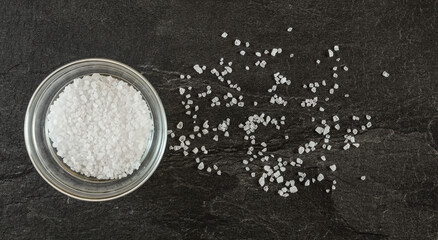 Small glass bowl with white rock salt on black marble like table - some crystals scattered near, view from above
