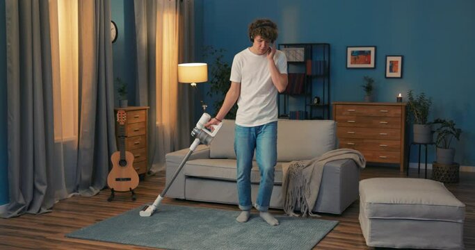 A crazy boy is cleaning the living room in the evening. The man is having a great time vacuuming the carpet in the room, listening to music on wireless headphones and dancing with the vacuum cleaner.