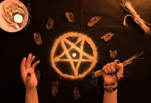 brujer&iacute;a, or&aacute;culo o lectura con runas de madera, estrella pentagonal de arena con manos de mujer sobre fondo negro, velas y amuletos
