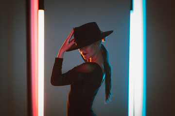 Stylish dancer woman in hat and bodysuit posing between neon led lamps on grey background. Unrecognizable lady looks hot. Art concept, femme fatale.