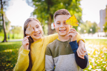 Happy twins teenagers boy and girl posing hugging each other in autumn park holding fallen yellow leaves in hand in sunny weather. Autumn season theme. Brother and sister have fun playing with leaves