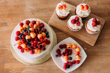 A cake with a large cap of berries (raspberries, cherries, blueberries) and buttercream. There are berries on the plate.  Four cupcakes with delicious cheese cream