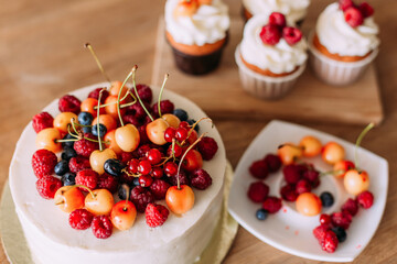 A cake with a large cap of berries (raspberries, cherries, blueberries) and buttercream. There are berries on the plate. Three cupcakes with delicious cheese cream