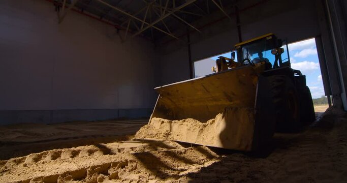 In a workshop under construction, a powerful wheeled tractor evens out the sand layer with its blade to create a cushion for the future floor 