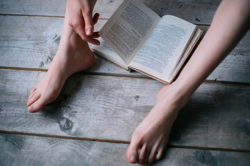A girl is reading a book barefoot, sitting on the floor. The book lies on the wooden floor between...