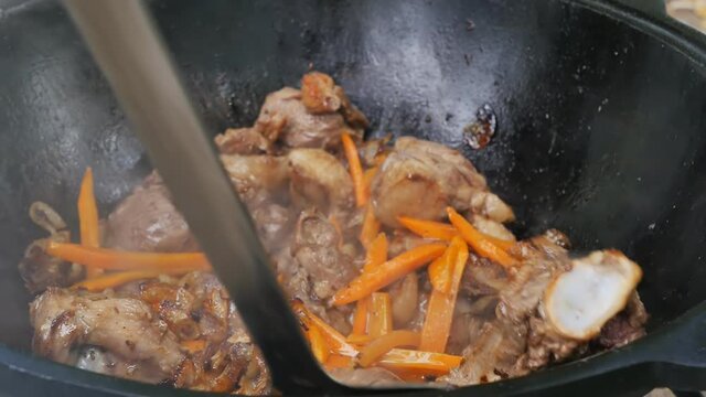 a slotted spoon mixes fried meat, carrots and onions in a large metal kettle. Cooking Uzbek pilaf. Close-up