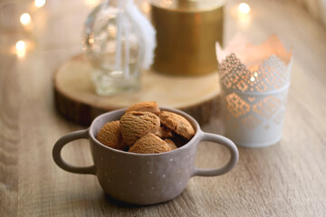 Bowl of cinnamon cookies, cup of warm drink, lit candles and vase with gypsophila flowers on the table. Selective focus, fairy lights in the background. Hygge at home.