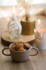 Bowl of cinnamon cookies, cup of warm drink, lit candles and vase with gypsophila flowers on the table. Selective focus, fairy lights in the background. Hygge at home.