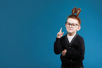Happy Easter kids. child boy in rabbit bunny ears on head with glasses on blue studio background. Pointing up. Looking and smiling at camera