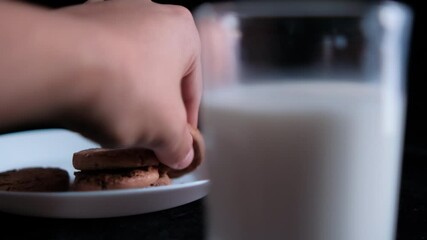 Hand grabbing cookie from a plate behind a glass of milk