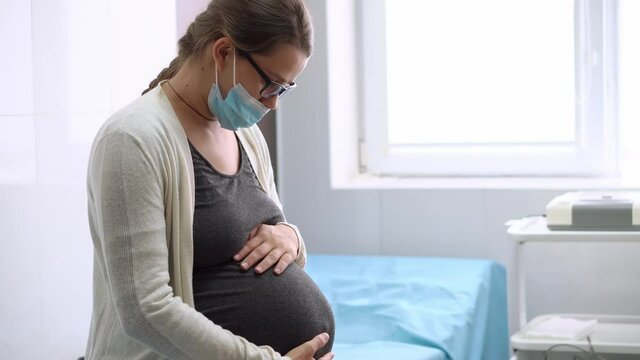 Young Pregnant Woman In Medicine Protective Mask On Hospital Couch Before Medical Control Cardiotocography. Visit Gynecologist Doctor At Clinic. Examine Expectant Belly Baby Mother Healthcare Check Up